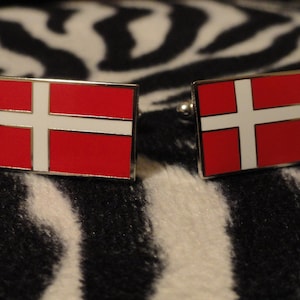 Könnte beinhalten: Ein Paar Manschettenknöpfe mit der dänischen Flagge. Die Manschettenknöpfe sind silberfarben und haben ein rotes und weißes Emaille-Design.