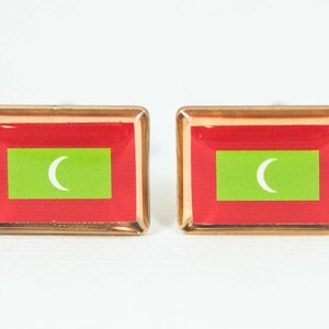 May include: A pair of cufflinks featuring the flag of the Maldives. The cufflinks are gold-toned and have a clear, glossy finish. The flag is red with a green rectangle in the center and a white crescent moon.