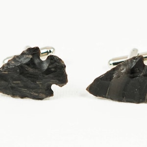 May include: A pair of cufflinks featuring a black arrowhead design. The arrowheads are made of a dark, textured material and are attached to silver cufflink backs.