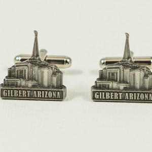 May include: Pair of silver-toned cufflinks featuring a detailed architectural design of a building with a spire. The base of each cufflink reads "GILBERT ARIZONA" in capital letters. The cufflinks have a classic, polished finish.