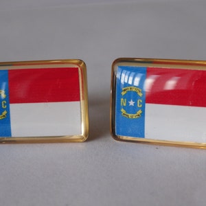 K&ouml;nnte beinhalten: Ein Paar goldene Manschettenkn&ouml;pfe mit der Flagge des Bundesstaates North Carolina. Die Flagge ist blau, wei&szlig; und rot mit einem Stern und den Buchstaben "N.C." im blauen Bereich.