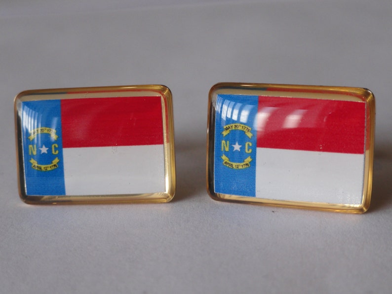 K&ouml;nnte beinhalten: Ein Paar goldene Manschettenkn&ouml;pfe mit der Flagge des Bundesstaates North Carolina. Die Flagge ist blau, wei&szlig; und rot mit einem Stern und den Buchstaben "N.C." im blauen Bereich.