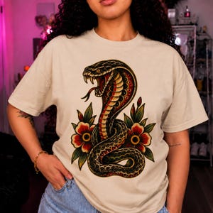May include: A beige short-sleeve t-shirt featuring a large graphic print of a coiled cobra snake with its mouth open, displaying fangs. The snake is surrounded by red flowers with yellow centres and green leaves in a traditional tattoo art style.