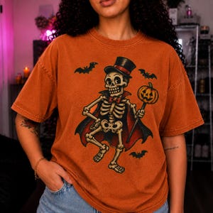 Puede incluir: Una camiseta de color naranja quemado con un gráfico de un esqueleto vestido de vampiro con sombrero de copa y capa, sosteniendo una calabaza en un palo. El diseño incluye tres pequeños murciélagos negros volando alrededor del esqueleto.