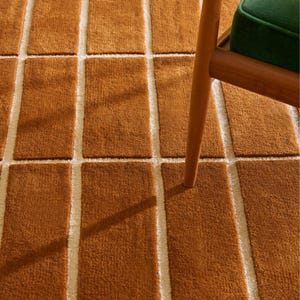 May include: Close-up of a brown rectangular rug with a grid pattern. The rug has a textured, plush appearance with off-white lines separating the brown rectangles. A wooden chair leg and a green cushion are visible in the upper right corner.