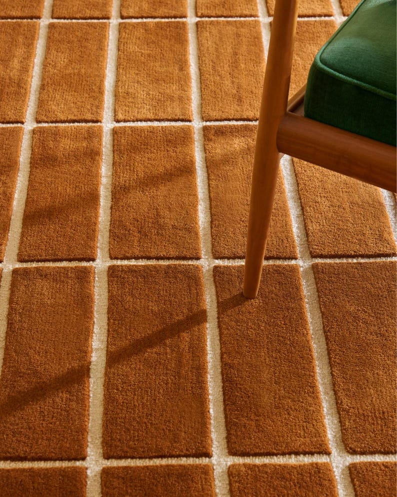 May include: Close-up of a brown rectangular rug with a grid pattern. The rug has a textured, plush appearance with off-white lines separating the brown rectangles. A wooden chair leg and a green cushion are visible in the upper right corner.