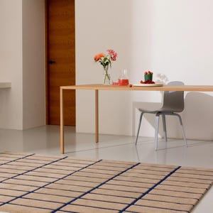 May include: A beige rectangular rug with a grid pattern of dark blue lines. The rug is on a light-colored floor, with a wooden table and a gray chair in the background. A vase of flowers and a small plate of berries are on the table.