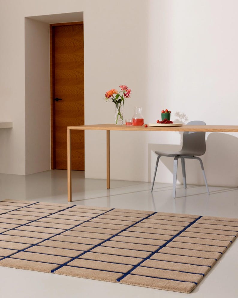 May include: A beige rectangular rug with a grid pattern of dark blue lines. The rug is on a light-colored floor, with a wooden table and a gray chair in the background. A vase of flowers and a small plate of berries are on the table.