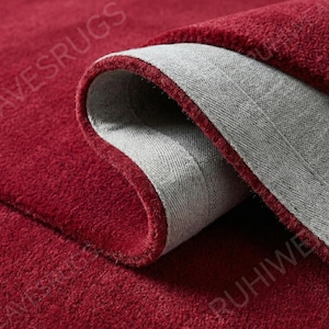 May include: Close-up of a rolled-up, plush red rug with a gray backing. The rug's edge is visible, showcasing its thick, soft texture. The rug's surface is a deep, solid red color.