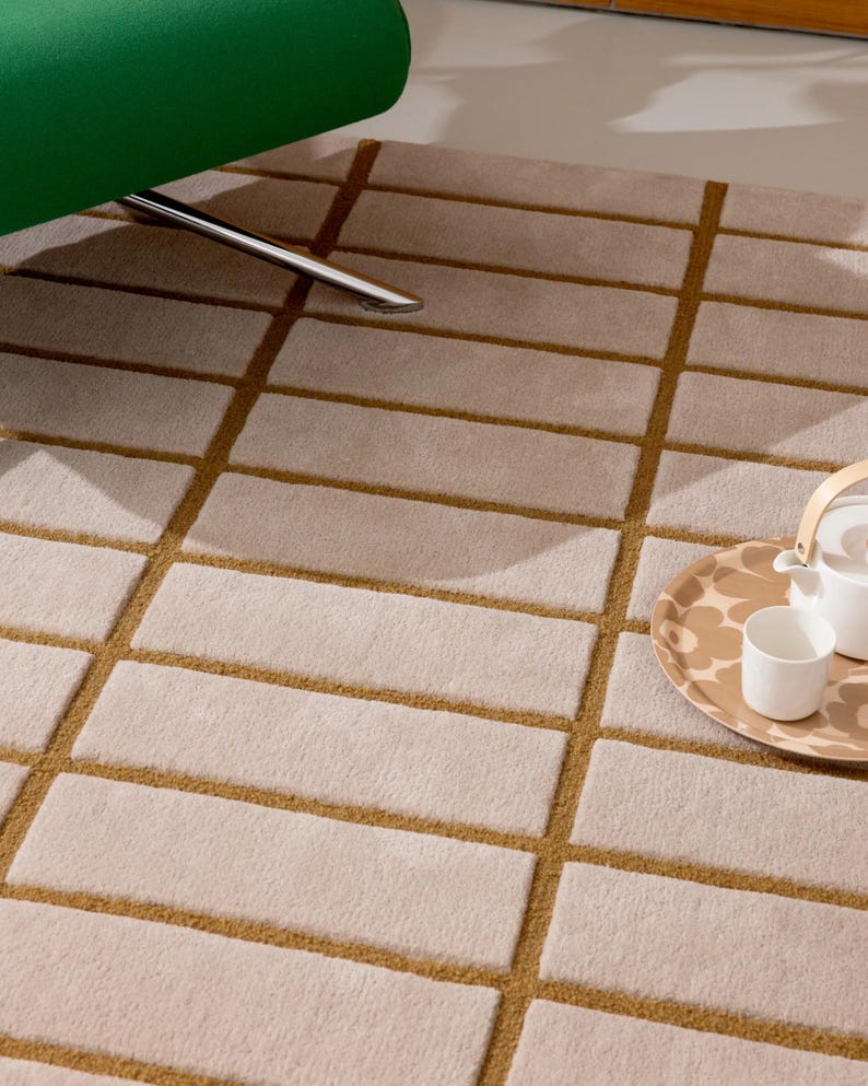 May include: A beige rectangular rug with a geometric pattern of light beige rectangles outlined in a darker tan color. A green chair leg is visible in the upper left corner, and a tray with a teapot and cup is in the right corner.