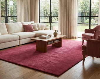 May include: A large rectangular area rug in a deep magenta hue, positioned on a light-toned wooden floor. The rug serves as a central foundation for a living room arrangement featuring a cream-colored sectional sofa and two matching magenta armchairs.