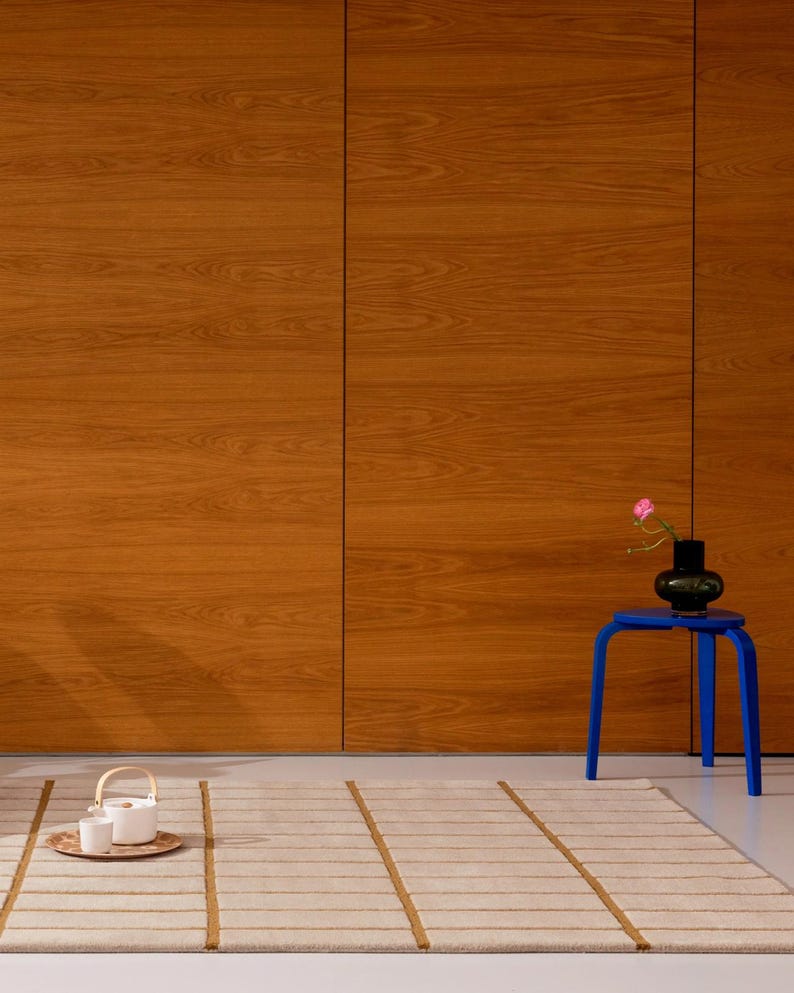 May include: A room scene featuring a light beige rug with brown stripes, a small blue table holding a black vase with a pink flower, and a wooden cabinet. A tea set sits on a wooden tray on the floor.