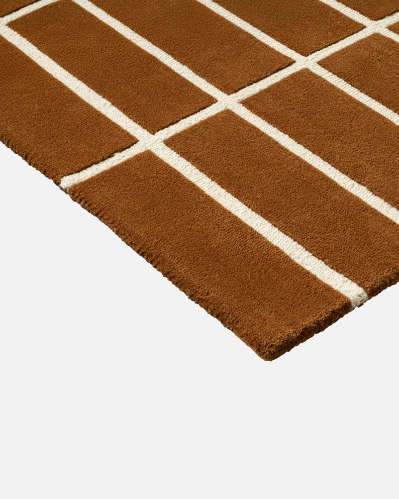 May include: Close-up of a brown rug with a geometric pattern. The rug features a grid design with thick, cream-colored lines against a brown background. The rug's texture appears soft and plush, suitable for home decor.
