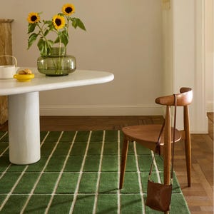 May include: A green rectangular rug with white lines, a white round table, and a wooden chair. A vase of sunflowers sits on the table. A brown leather bag hangs on the chair.