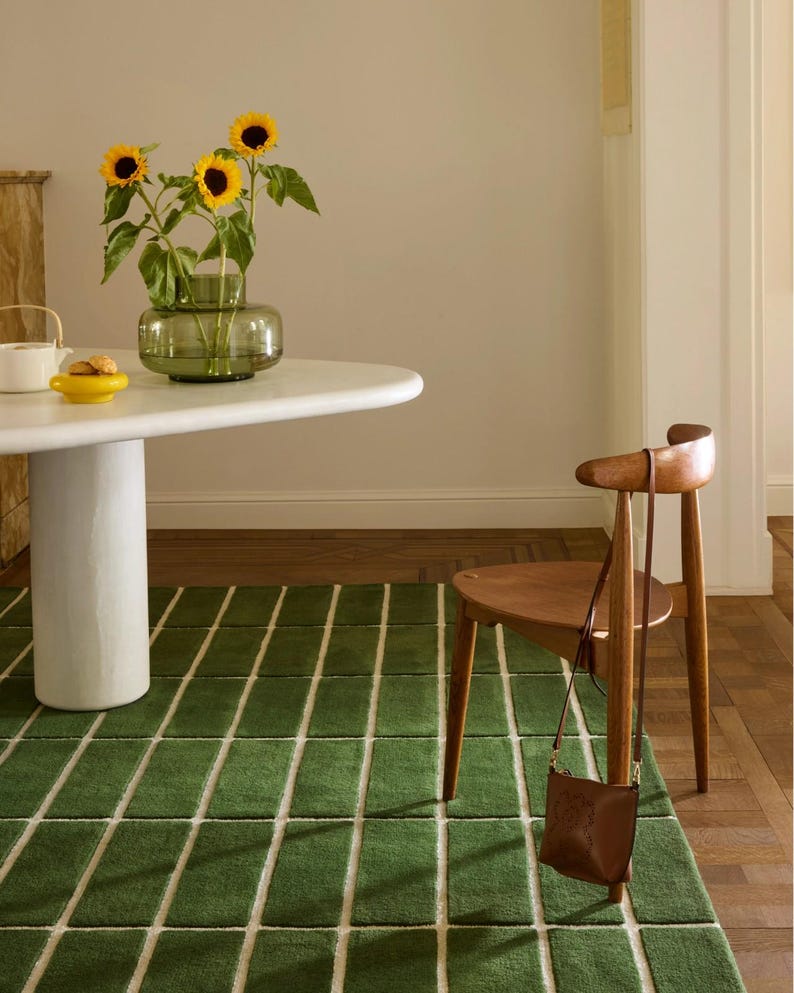 May include: A green rectangular rug with white lines, a white round table, and a wooden chair. A vase of sunflowers sits on the table. A brown leather bag hangs on the chair.