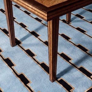 May include: A wooden table with a polished, light brown burl wood top and dark brown edges, featuring four straight, vertical legs. The table is positioned on a light blue rug with a repeating black and tan geometric step pattern.