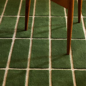 May include: A green rectangular rug with a grid pattern of off-white lines. The rug is made of a soft, textured material. The legs of a wooden chair are visible in the image.