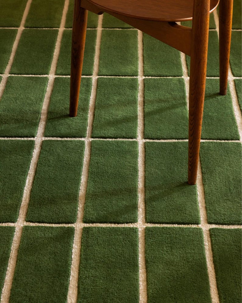 May include: A green rectangular rug with a grid pattern of off-white lines. The rug is made of a soft, textured material. The legs of a wooden chair are visible in the image.