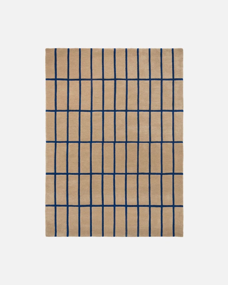 May include: A rectangular beige rug with a grid pattern of dark blue lines. The lines are evenly spaced, creating a modern and geometric design. The rug is made of a soft, textured material.