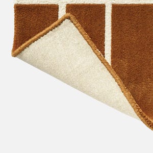 May include: Close-up of a brown rug with a grid pattern in white. The rug's corner is lifted, revealing a beige backing and a brown border. The rug has a soft, textured appearance.