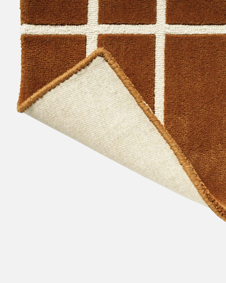 May include: Close-up of a brown rug with a grid pattern in white. The rug's corner is lifted, revealing a beige backing and a brown border. The rug has a soft, textured appearance.