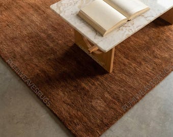 May include: A brown rug with a textured surface, paired with a marble-topped coffee table and an open book. The rug is a warm, earthy tone, and the table has wooden legs. The text "RUHIWEAVESRUGS" is visible on the rug.