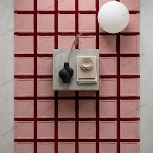 Pode incluir: Um tapete retangular rosa com um padrão de grade vermelho escuro. Uma pequena mesa quadrada com um vaso preto e livros está sobre o tapete. Uma luminária branca em forma de esfera está ao fundo. Mede aproximadamente 200 cm x 300 cm.