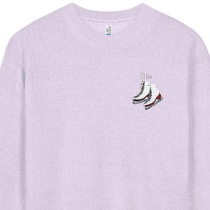 May include: A light purple crewneck sweatshirt featuring a small graphic on the upper left chest. The graphic depicts a pair of white ice skates with red accents and silver blades, accompanied by the text "I Love" written in a thin, black script.