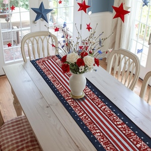 May include: A rectangular table runner featuring a patriotic design with red, white, and blue stripes, stars, and intricate patterns. The runner is centered on a wooden dining table, accompanied by a white vase filled with red and white flowers.