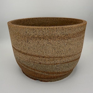 May include: A round ceramic planter with a textured, speckled surface in shades of tan, beige, and brown. The exterior features horizontal, swirling bands of darker earth tones that wrap around the body of the vessel, creating a natural, grainy appearance with a wide, open top rim.