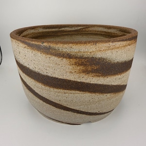 May include: A round ceramic planter with a speckled beige and tan base featuring wide, swirling dark brown stripes. The vessel has a thick, textured rim and a wide opening, showcasing a natural, earthy clay finish with visible granular details.