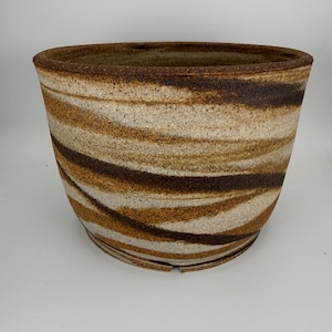 May include: A wide, tapered ceramic planter featuring a textured, speckled surface with swirling horizontal bands of earthy brown, tan, and cream tones. The rim is thick and dark, and the base has a small notch visible at the bottom edge.