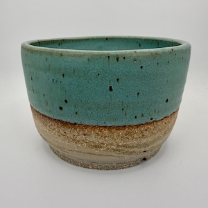 Op de afbeelding: Een handgemaakte keramische kom met een turquoise glazuur op de bovenste helft en een natuurlijke, getextureerde bruine afwerking op de onderste helft. De kom heeft een breed, open ontwerp, geschikt om te serveren of tentoon te stellen. De kom heeft een diameter van ongeveer 15 cm.