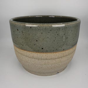May include: A ceramic planter featuring a two-tone design with a glossy, speckled sage green glaze on the upper portion and a textured, matte beige finish on the lower section. The bowl-shaped vessel has a wide opening and a rounded base.