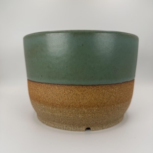 May include: A two-toned ceramic planter. The top half is a matte teal green, and the bottom half is a textured, earthy brown. The planter has a simple, rounded shape, suitable for various plants. The design is minimalist and modern.
