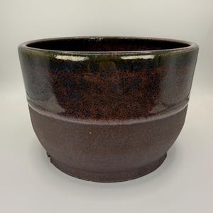Peut inclure: Un pot de fleurs en céramique au design bicolore, composé d'une partie supérieure émaillée lisse, brillante, brun foncé et violette, et d'une base mate et texturée brun foncé. Ce récipient en forme de bol possède une large ouverture.