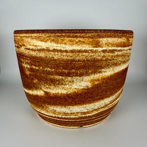 May include: A handcrafted, wide-mouthed ceramic bowl with a unique, layered design. The bowl features alternating bands of brown and cream colors, creating a textured, earthy aesthetic. The bowl's shape is slightly tapered, with a flat base.