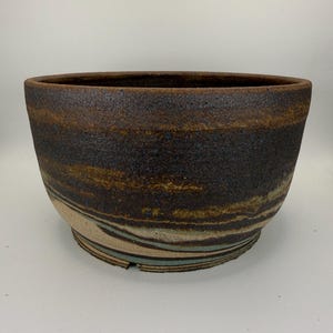 May include: A wide, round ceramic bowl with a textured, dark brown and black exterior featuring horizontal bands of tan and muted blue-green marbling near the base. The rim is smooth and dark, contrasting with the rough, earthy finish.