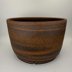 May include: A wide, round ceramic planter with a textured, dark brown and burnt orange mottled finish. The exterior features horizontal bands of varying earthy tones and a thick, rounded rim. The base is flat with a small notch visible at the bottom.