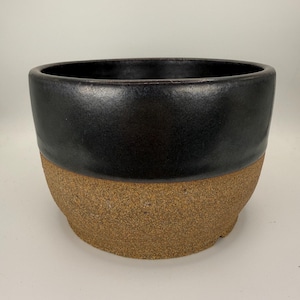 May include: A round ceramic planter featuring a two-tone design. The upper portion has a smooth, glossy black glaze, while the lower portion displays a textured, speckled brown clay finish. The vessel has a wide, open top and a sturdy base.