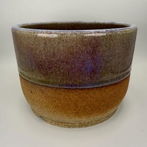 Peut inclure: Un pot de fleurs cylindrique en céramique avec une finition bicolore. La partie supérieure présente un vernis brillant moucheté de violet et de gris, tandis que la section inférieure affiche une surface en terre cuite brune, mate et granuleuse.