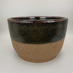 Puede incluir: Un cuenco de cerámica con un diseño de dos tonos, que presenta una sección superior esmaltada en verde oliva oscuro y una base texturizada de color tostado con motas. El cuenco tiene un borde circular ancho y un cuerpo redondeado hacia la base.