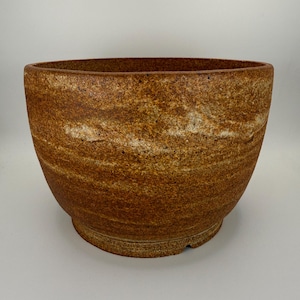 May include: A wide, round ceramic bowl with a textured, mottled surface in shades of earthy brown, tan, and cream. The vessel features a thick, rounded rim and a slightly tapered base, showcasing a granular, speckled finish throughout the exterior.