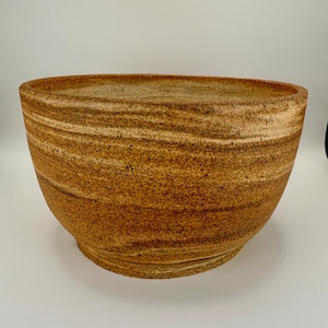 May include: A large, handcrafted ceramic bowl with a wide, oval opening. The bowl features horizontal bands of tan and brown hues, creating a textured, layered appearance. The base is a slightly darker shade of brown. This decorative bowl is suitable for home decor.