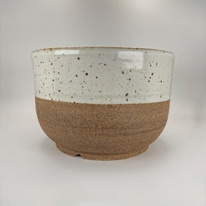 May include: A handcrafted ceramic bowl with a two-tone design. The upper half is a speckled off-white, while the lower half is a textured brown. The bowl has a rounded shape and a wide opening, suitable for serving or display.
