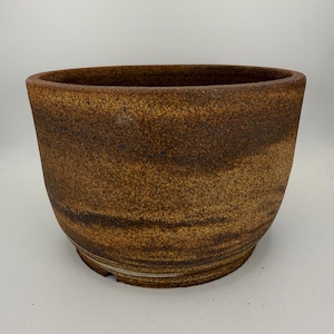 May include: A round ceramic planter with a textured, speckled surface in shades of deep brown, tan, and ochre. The vessel features a wide, open top and a slightly tapered base with a visible horizontal ridge near the bottom edge of the exterior.