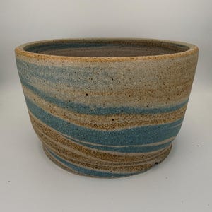 May include: A wide, round ceramic planter featuring a marbled pattern of tan, beige, and blue swirls. The exterior has a speckled, textured finish with a wide opening and a slightly tapered base, showcasing a handcrafted clay construction.
