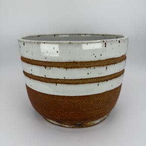 May include: A handmade ceramic bowl with a speckled white rim and two horizontal brown bands. The lower portion of the bowl is a textured brown, creating a rustic and earthy aesthetic. The bowl is suitable for serving food.