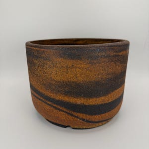 May include: A round ceramic planter with a dark brown and orange swirl pattern. The planter has a wide opening and a textured surface. The colour palette is earthy and natural, suitable for indoor or outdoor use.
