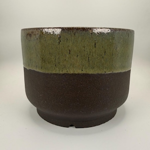 Puede incluir: Una maceta de cerámica de dos tonos con forma redondeada. La mitad superior es de color verde oliva brillante, mientras que la mitad inferior es de color marrón oscuro texturizado. La maceta tiene una abertura amplia y una base robusta, adecuada para varias plantas.